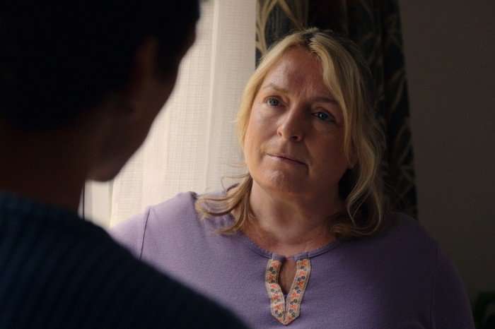 Claire Rushbrook as Dr Jean Harris-Hendriks in The Witness standing in a room by a curtain, looking seriously at someone while wearing a purple top.
