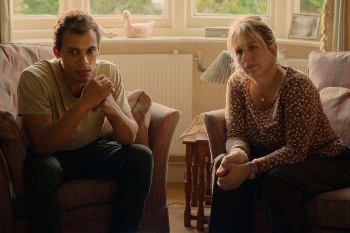 Jordan Bolger as Andr&eacute; Hanscombe and Kerry Godliman as June in The Witness sitting in armchairs in a living room, looking worried as the light comes in from behind them from the windows.