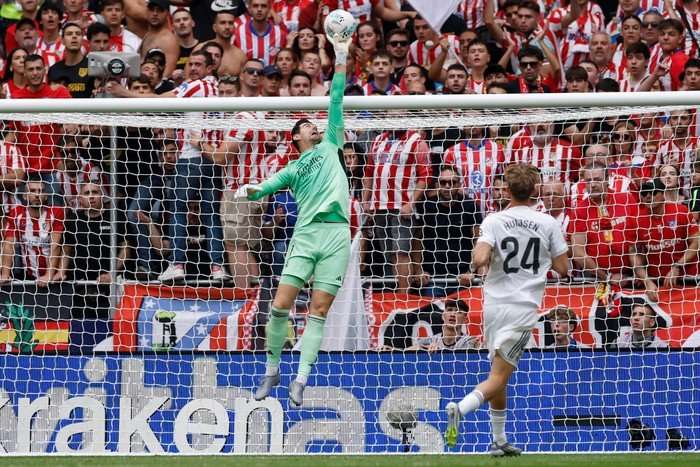 Thibaut Courtois, in light green Real Madrid goalkeeping kit and white gloves, tips a football over the crossbar with Atletico Madrid fans behind him.