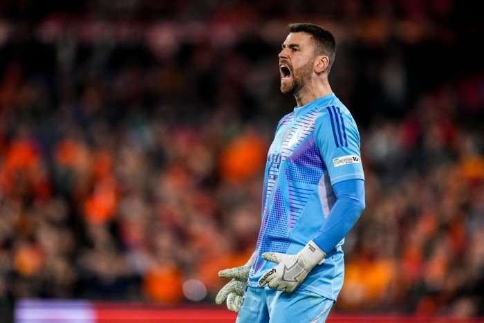 Unai Simon, in blue Spain goalkeeping kit and grey gloves, roars with Dutch fans blurred in the background.