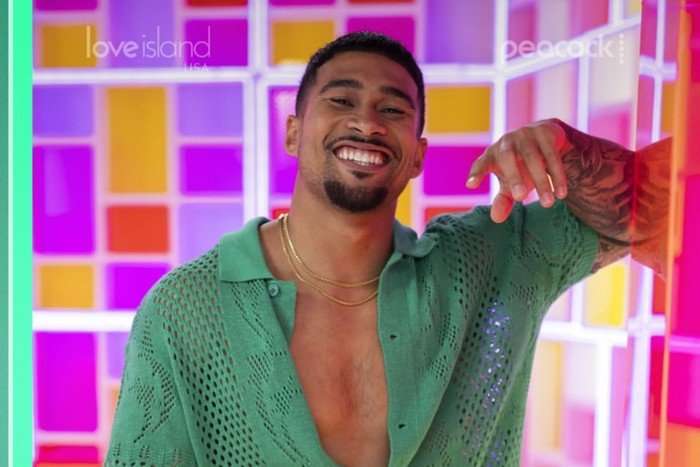 A close up photo of a brunette man standing in front of a geometric panel background wearing an unbuttoned green crochet shirt. He has one hand resting on a wall and the other beside him as he smiles ahead.