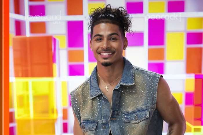 A close up photo of a brunette man standing in front of a geometric panel background wearing an unbuttoned denim sleeveless vest and smiles ahead.