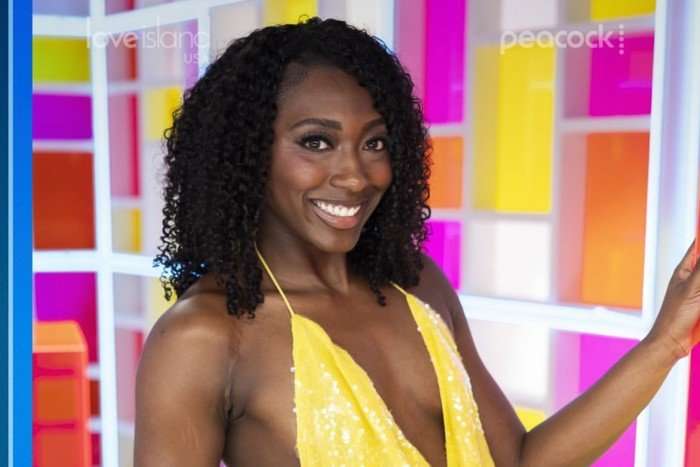 A close up photo of a brunette woman standing in front of a geometric panel background wearing a yellow sparkle dress. She has one hand resting on a wall and the other beside her as she smiles ahead.