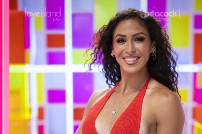 A close up photo of a brunette woman standing in front of a geometric panel background wearing a red dress and smiling ahead.