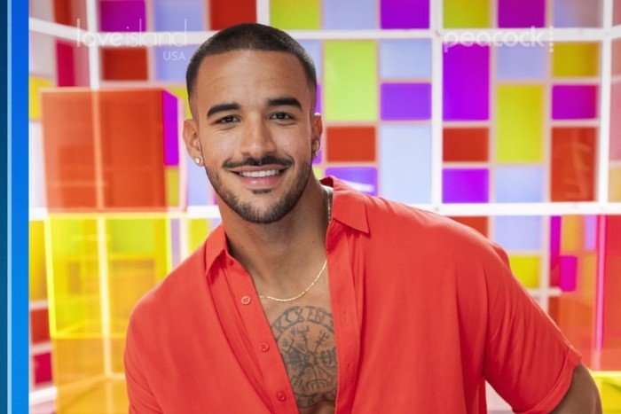 A close up photo of a brunette man with a tattooed chest wearing an unbuttoned red shirt and smiling ahead.
