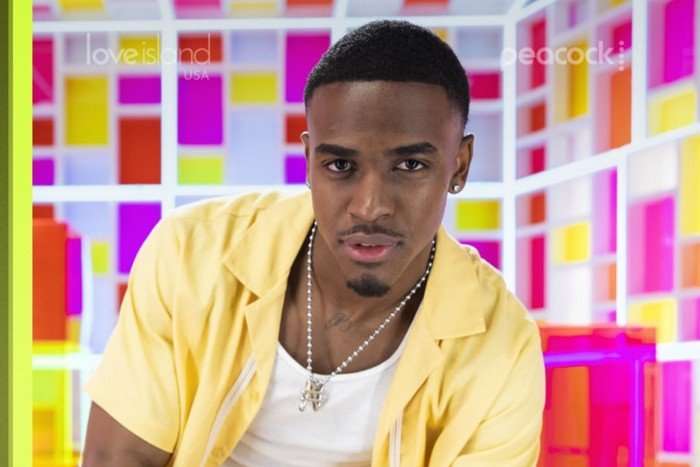 A close up photo of a brunette man standing in front of a geometric panel background wearing an unbuttoned yellow shirt with a white vest underneath looking ahead.