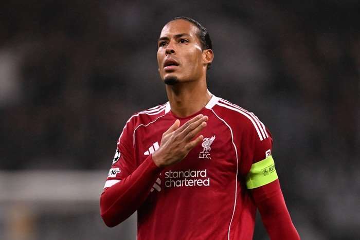 Virgil van Dijk Virgil van Dijk, in red Liverpool football kit, raises his right hand to the club badge as he looks up to the crowd.