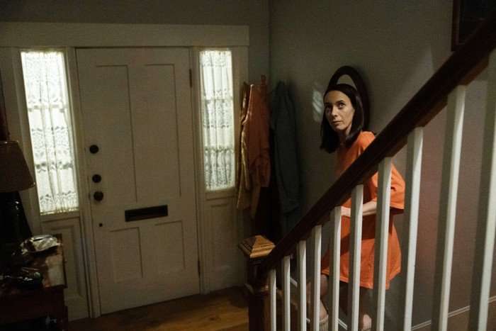 Kate O'Flynn as Patricia in Widow's Bay episode 8. She's cautiously walking downstairs in the interior of a house, wearing an oversized red Tshirt.