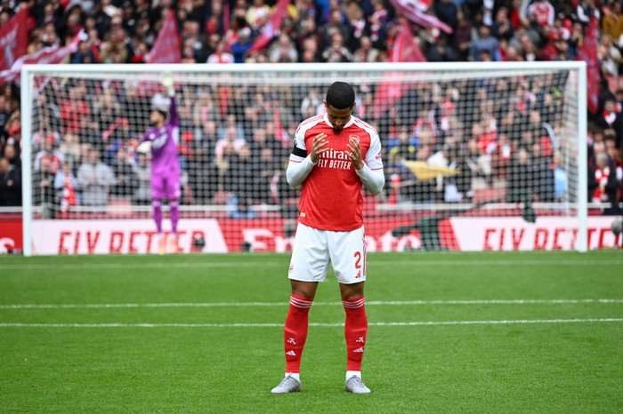 William Saliba William Saliba, in red and white Arsenal football kit, bows his head in prayer with the goal and crowd in the background.