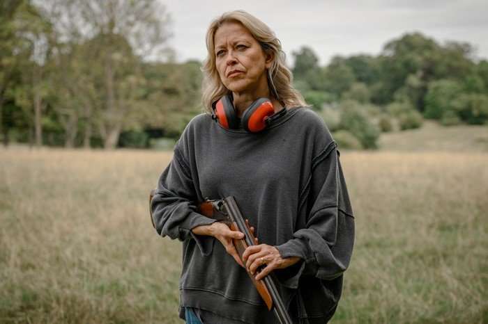 alice-steve-nicola-walker Alice holds a shotgun and stands in a field, looking unimpressed, with protective headphones resting around her neck