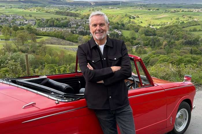 Brian Conley photographed in front of a red convertible car, with rolling green hills of the English countryside behind him.
