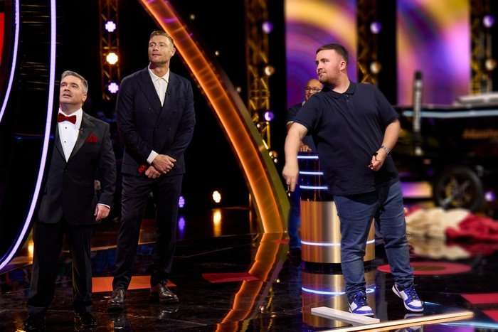 ©ITV Freddie Flintoff and a co-host watch as Luke Littler throws darts during a game on Bullseye's vibrant stage