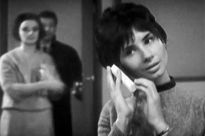 c001-susan-dancing-doctor-who-unearthly-child-whobackwhen.v1 A black and white image from Doctor Who featuring Susan, a young woman with short hair, holding a transistor radio to her ear, her expression thoughtful. In the softly focused background, a couple, Ian and Barbara, stands close together.