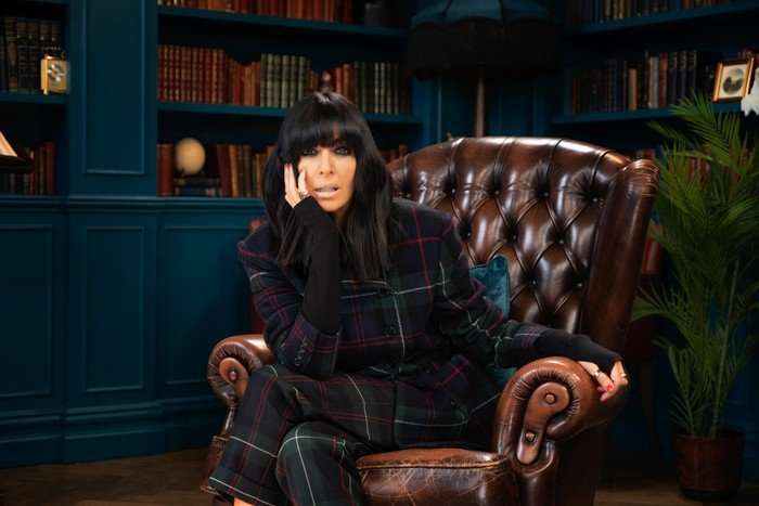 Claudia Winkleman sitting in a red armchair in a study with her hand to her face on the set of The Traitors.