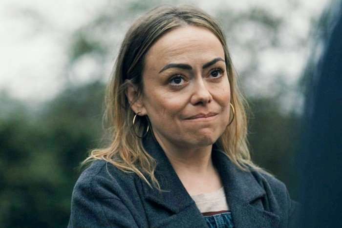 A close-up of a woman outdoors with a thoughtful, slightly tense expression. She has shoulder-length light brown hair parted in the middle, dark eyebrows, and is wearing large hoop earrings, a dark coat, and a light-colored top. The background is blurred, showing trees and an overcast sky, while part of another person is visible out of focus in the foreground on the right.