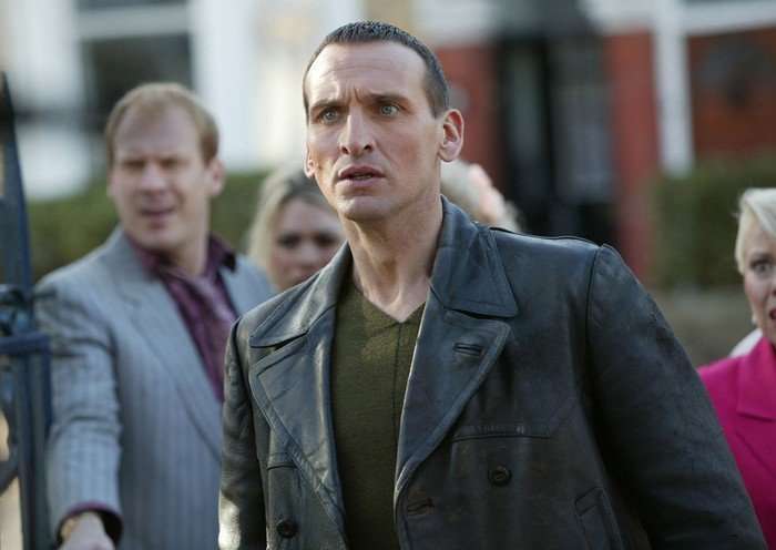 Christopher Eccleston as the Ninth Doctor in Doctor Who, wearing his iconic black leather jacket and green jumper, stands outdoors looking tense, with several people in the background also appearing alarmed.