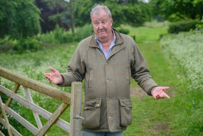 clarksons-farm (2) Jeremy Clarkson wearing a blue shirt and green parka jacket. He is stood in the middle of grass with his arms stretched out as he shrugs.