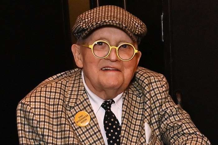 david-hockney David Hockney wearing a printed suit and cap, smiling as she looks ahead.