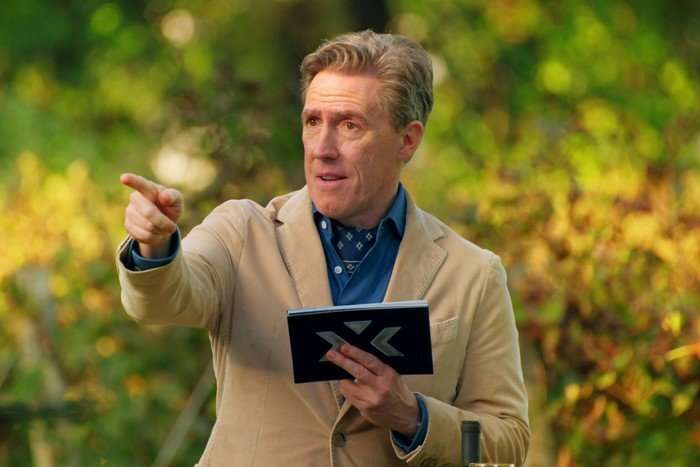 Rob Brydon holding a set of cards in one hand and pointing at something with the other as he stands in a grassy field.
