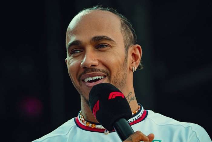 drive-to-survive Lewis Hamilton wearing a white t-shirt and holding a microphone and smiling.