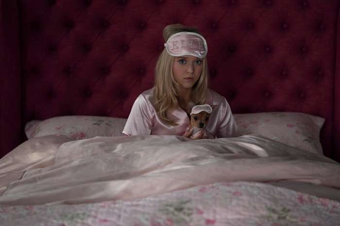 Lexi Minetree as Elle Woods in Elle sitting in bed, wearing pink satin pyjamas and an eye mask with her chihuahua in her lap in a matching Elle mask.