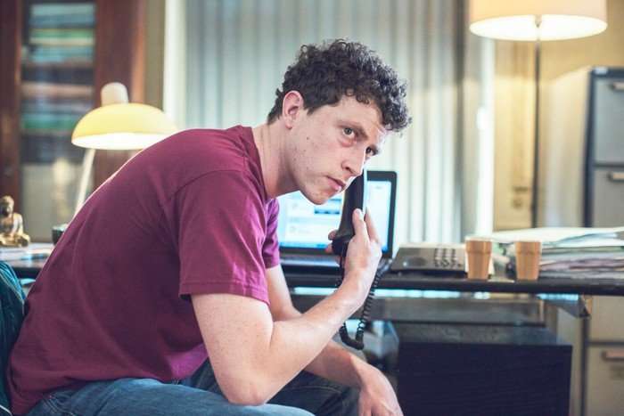 GONE FOR GOOD SEASON 1 A young man in a burgundy T-shirt sits at a desk in a softly lit room, holding a landline telephone receiver to his ear. He looks off to one side with a tense, concerned expression. Behind him, an open laptop, a desk phone, stacks of papers and disposable coffee cups suggest an ongoing investigation or urgent work. Warm lamp light and muted tones create a serious, suspenseful atmosphere.