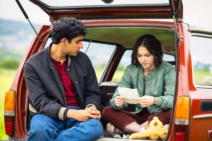 A Good Girl's Guide To Murder Ravi and Pip sat in the open boot of a car, looking at a piece of paper