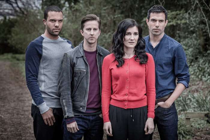 The Five Four adults stand together on a wooded path, facing forward with serious, concerned expressions. A woman in a bright red cardigan stands at the front, flanked by three men wearing casual clothing, including a leather jacket and a blue button-up shirt. Trees and dense greenery fill the background, creating a tense, dramatic atmosphere.