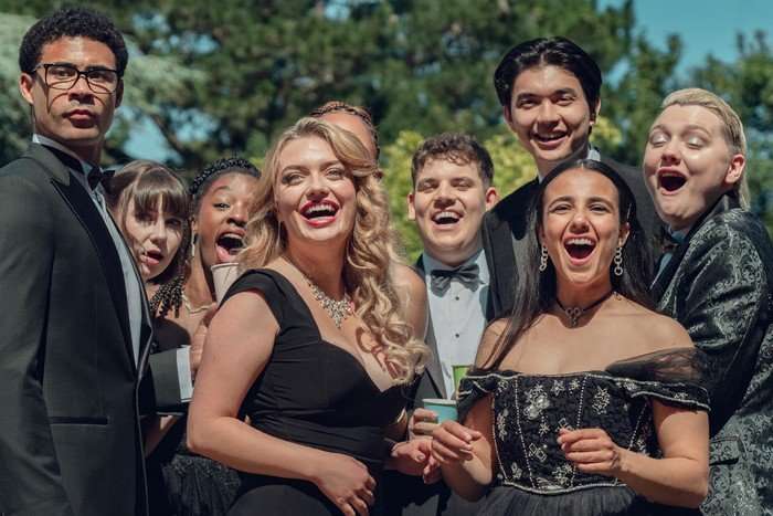 heartstopper-forever-group The main characters of Heartstopper wear formal black tie outfits and stand together in a garden. They are laughing in delight at something in the distance ahead of them.