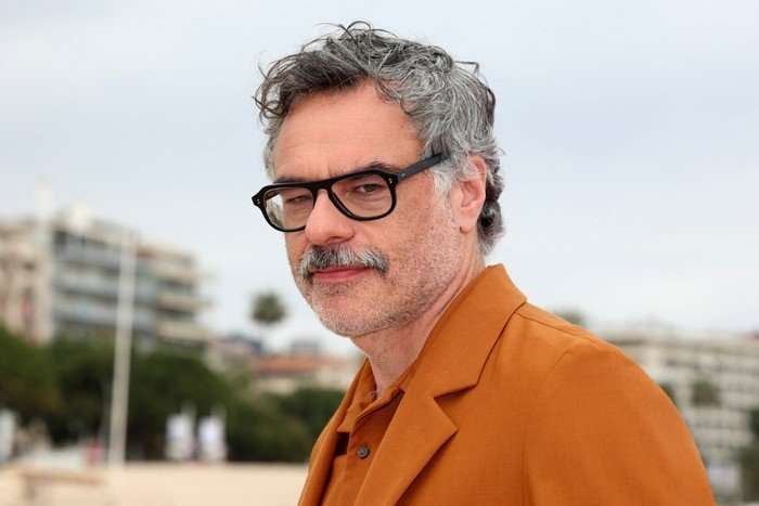 Close-up of Jemaine Clement wearing an orange suit and looking towards the camera.