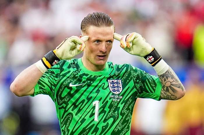 Jordan Pickford wearing the bright green England goalkeeping kit and white gloves. He is pointing to his head with both hands.