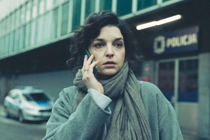 Just One Look A woman with dark curly hair stands outside a police station, holding a mobile phone to her ear. Wrapped in a grey coat and scarf, she looks anxious and distracted as she listens to the call. A police car is parked in the background, while the station entrance and signage are visible behind her. The muted colours and overcast setting create a tense, suspenseful mood.