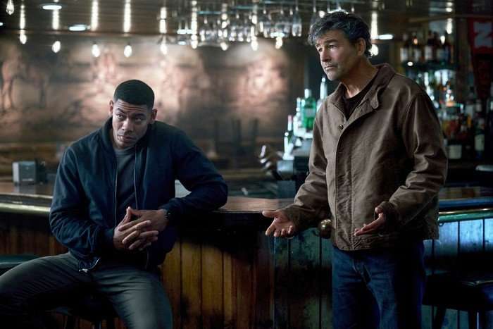 lanterns-aaron-pierre-kyle-chandler-3 Aaron Pierre and Kyle Chandler as John Stewart and Hal Jordan in Lanterns. Their characters are in a bar decorated with white fairy lights, speaking to someone as part of their criminal investigation.