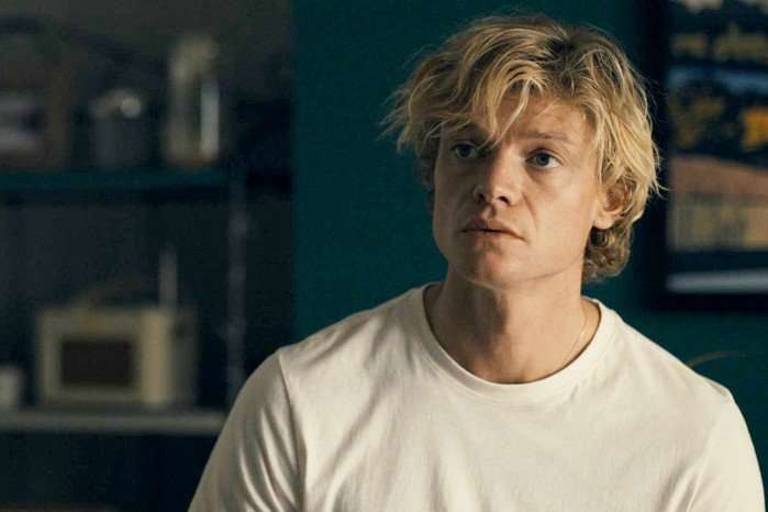 A young person with tousled blond hair and a serious, attentive expression sits indoors, looking slightly off-camera. They are wearing a plain white T-shirt and are lit by warm indoor lighting. The background is softly out of focus, showing shelving and household items against a dark teal wall, creating an intimate, conversational atmosphere.
