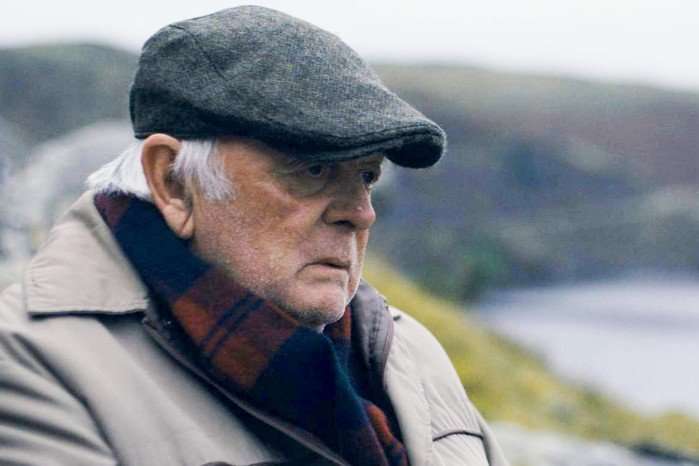 An elderly man wearing a grey flat cap, beige coat, and a red-and-navy checked scarf sits outdoors near a body of water. He gazes into the distance with a thoughtful, somber expression. The background is softly blurred, showing a shoreline and hills under an overcast sky, creating a quiet and reflective mood.