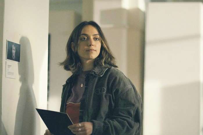 A young woman with shoulder-length dark hair stands indoors holding a folder or clipboard while looking off to one side. She wears an oversized dark jacket over a burgundy top and has a calm, observant expression. Beside her on the wall is a small display featuring a photograph and text, suggesting a gallery, exhibition, or public building. Soft, natural light fills the modern interior, and her shadow is cast clearly on the wall behind her.