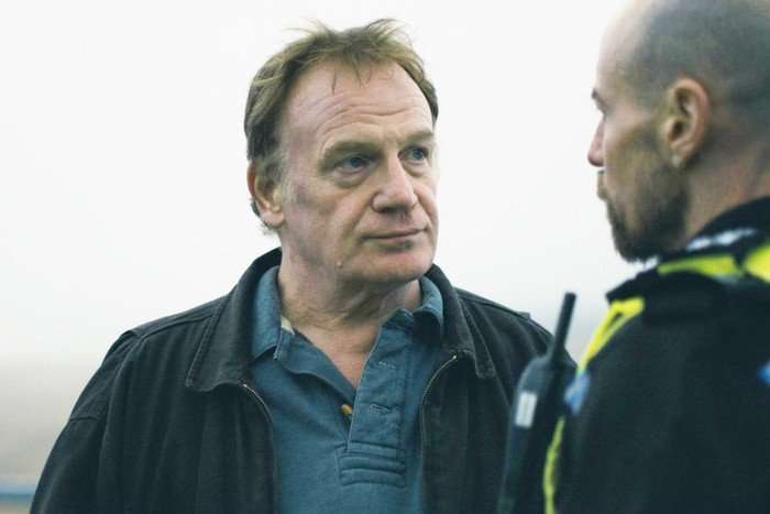 A middle-aged man in a dark jacket and blue collared shirt looks intently at a police officer standing in front of him. The officer is shown in profile and slightly out of focus, wearing a high-visibility uniform with a radio attached. The scene appears to be outdoors on an overcast day, with a pale, blurred background that emphasizes the serious exchange between the two men.