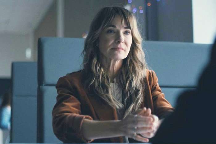 A woman with long wavy brown hair and bangs sits at a table in a softly lit indoor setting, facing someone off-camera. She wears a rust-coloured blazer over a light top and has her hands clasped in front of her. Her expression is calm and attentive, suggesting a serious conversation. The background is softly blurred, with upholstered seating and small decorative lights visible behind her.