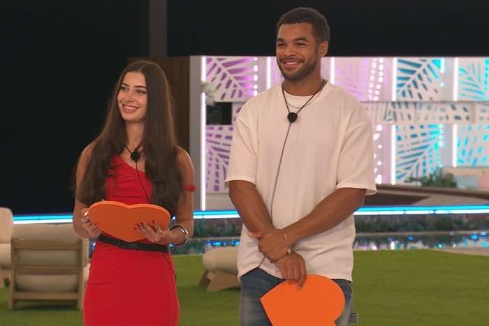 love-island - 2025-08-01T100733.457 Yasmin and Jamie stood next to each other, smiling ahead as they hold orange boards shaped as hearts.