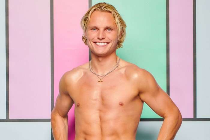 A topless man posing with his hands on his hips, smiling ahead in front of a colour block background.