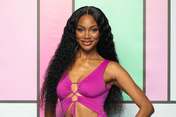 A woman wearing a pink swimsuit, smiling ahead, with one hand on her hip.