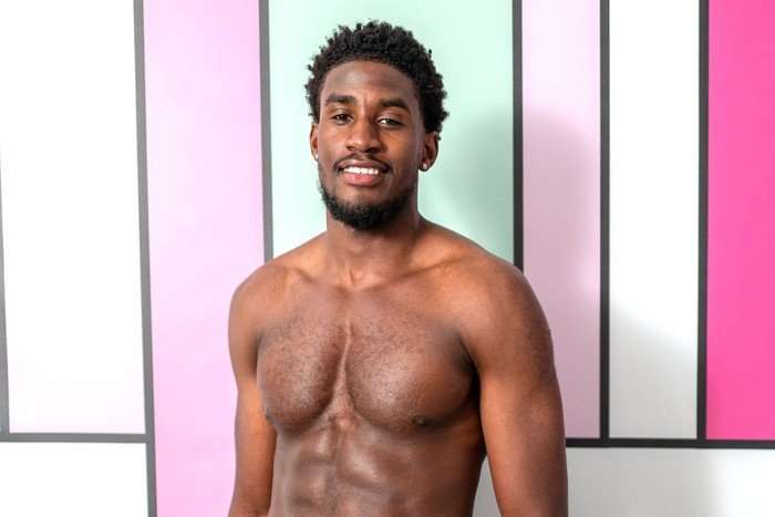 love-island (45) A topless man smiling ahead stood in front of a pastel blue and pink background.