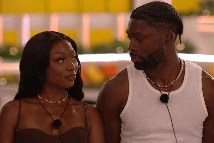 love-island-cast Mimii and Josh looking at each other, both smiling. Mimii is wearing a brown dress with mic around her neck while Josh is wearing a white vest and also has a mic around his neck.