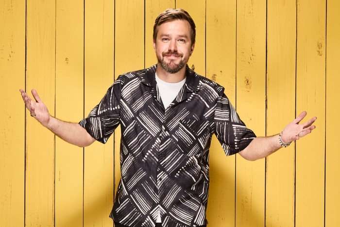 Iain Stirling posing with his arms stretched out, wearing a black and white shirt in front of a yellow wooden background.