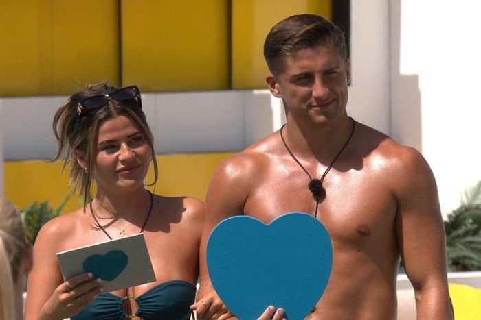love-island-recap-46 (1) Matilda and Sean stood next to each other in the villa garden, holding a blue heart and cue cards. They are looking across and smiling.