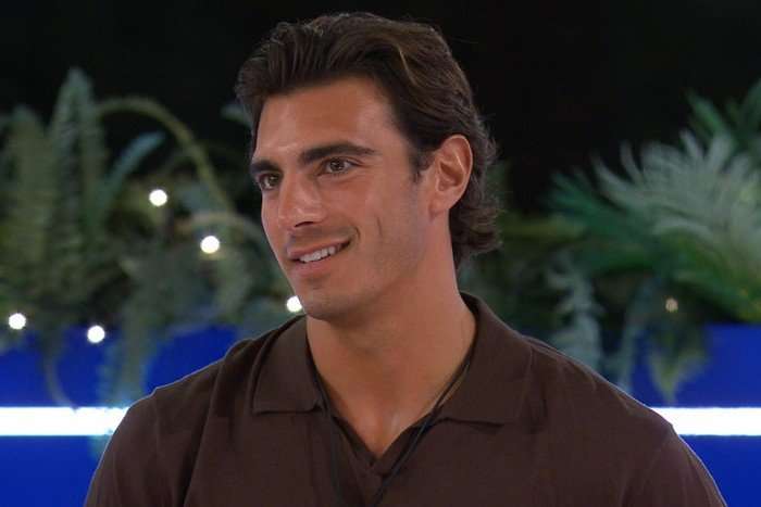 Love Island S13 Lorenzo stood up at the fire pit smiling ahead as he looks at someone.