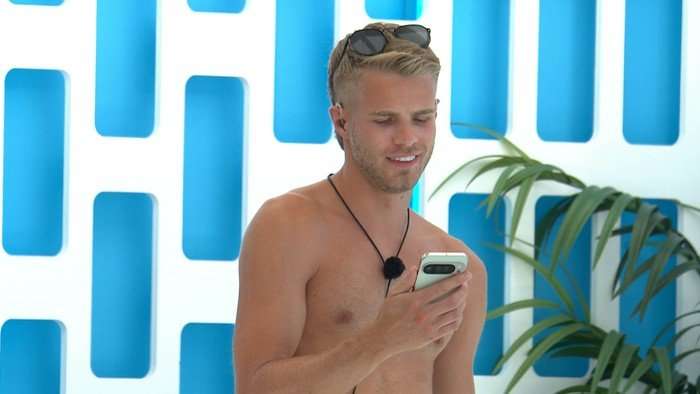 Love Island star Sam shirtless with sunglasses resting on his head smiles while looking at a smartphone, standing indoors against a blue-and-white decorative wall with a leafy plant in the background
