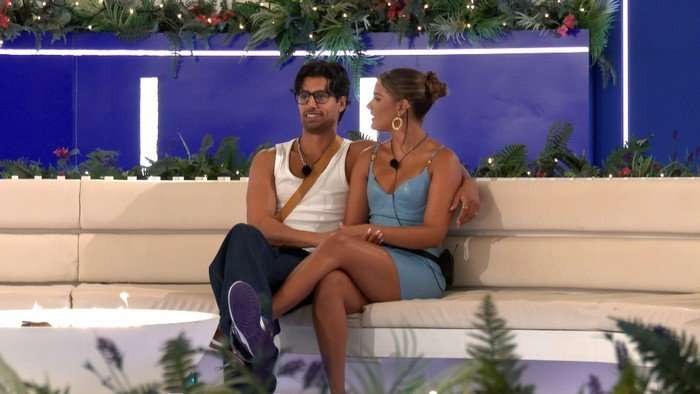 Mica looking at Samraj at the fire pit, who has his arm round her, both smiling.