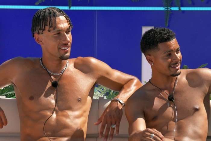 Jordon and Carlos sat down next to each other looking over at people across from them. They are both topless and are resting their arms behind them.