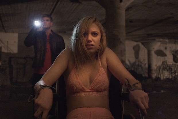 maika_monroe_it_follows Maika Monroe stars in It Follows sitting in a chair with her hands tied, with a man holding a torch in the background